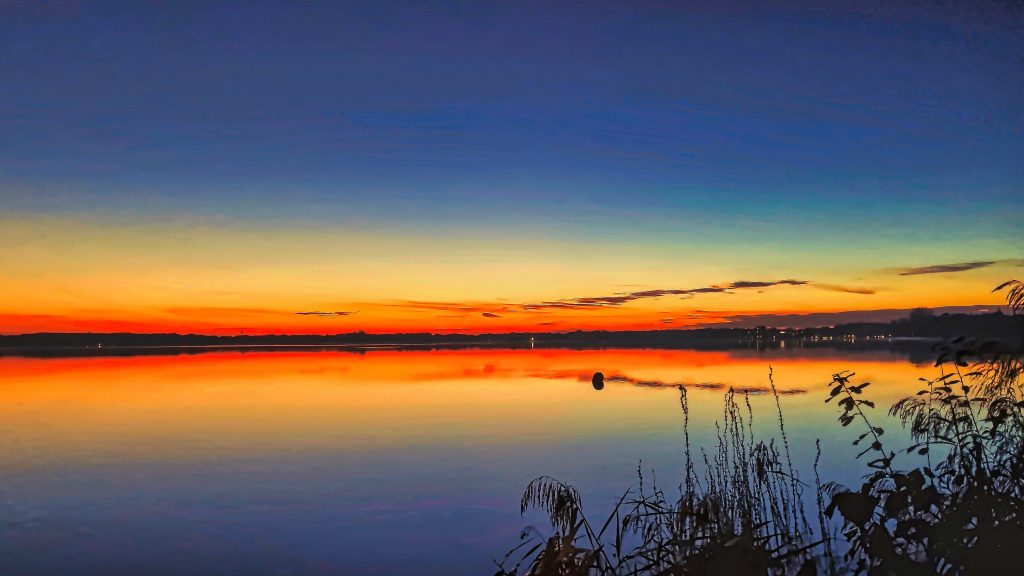 Landschaftsfotografie eines farbenreichen Sonnenuntergangs über einem ruhigen See mit Spiegelung und Silhouetten im Vordergrund.