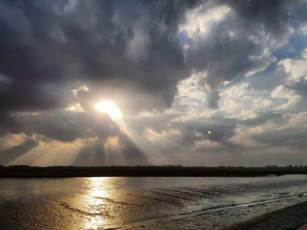 Dramatischer Himmel über einem Wattgebiet bei Sonnenuntergang. Sonnenstrahlen brechen durch dunkle Wolken und spiegeln sich im nassen Sand des Wattenmeeres. Im Hintergrund sind Windräder und eine weite Landschaft zu erkennen.