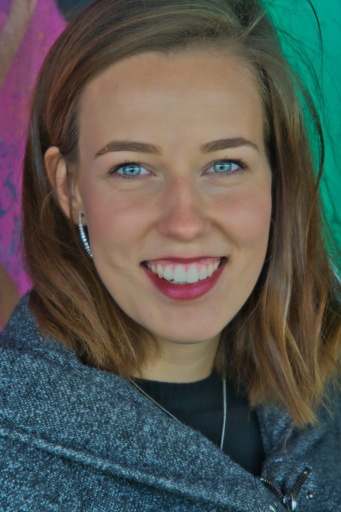 Porträt einer lächelnden jungen Frau mit blauen Augen und braunen Haaren vor farbenfrohem Hintergrund.