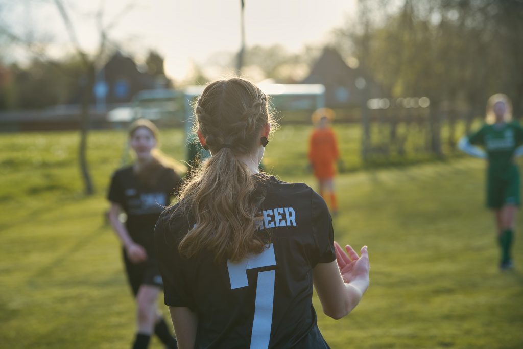 Fußballspielerin mit der Nummer 7 steht auf dem Spielfeld bei Sonnenuntergang, andere Spielerinnen im Hintergrund sichtbar. Passend zum Thema Sport Fotografie.