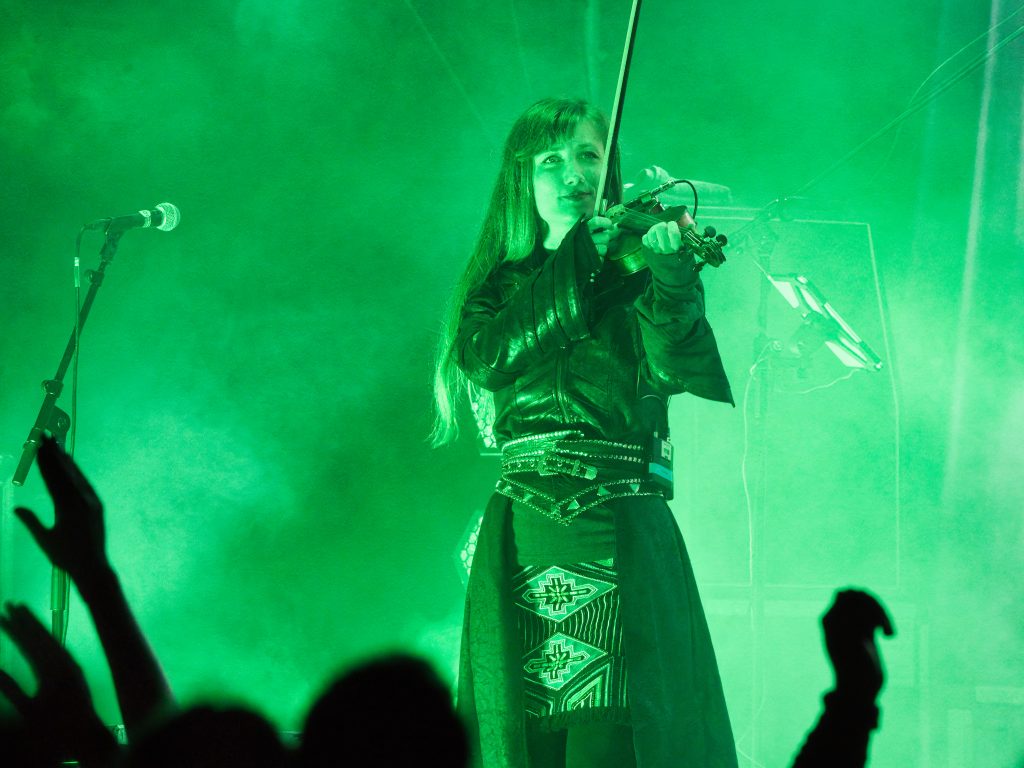 Geigerin spielt auf der Bühne im grünen Licht vor jubelndem Publikum. Sie ist Fantasy artig gekleidet.