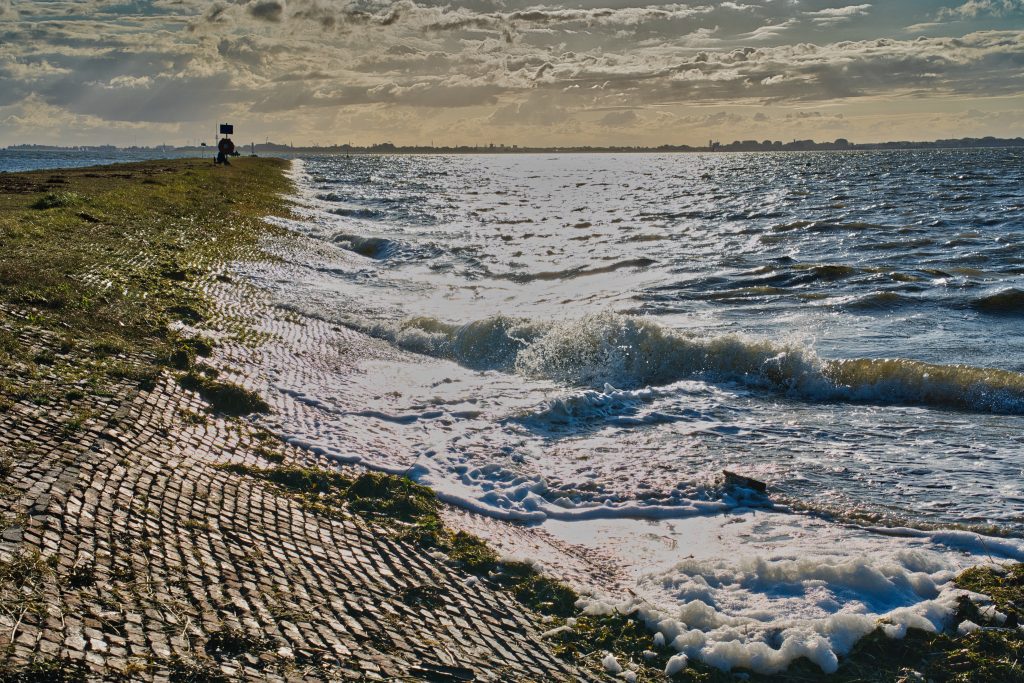 Wellen schlagen gegen den Deich an der Nordsee bei Sonnenschein und Wind, mit Schaum und Wolken am Horizont.