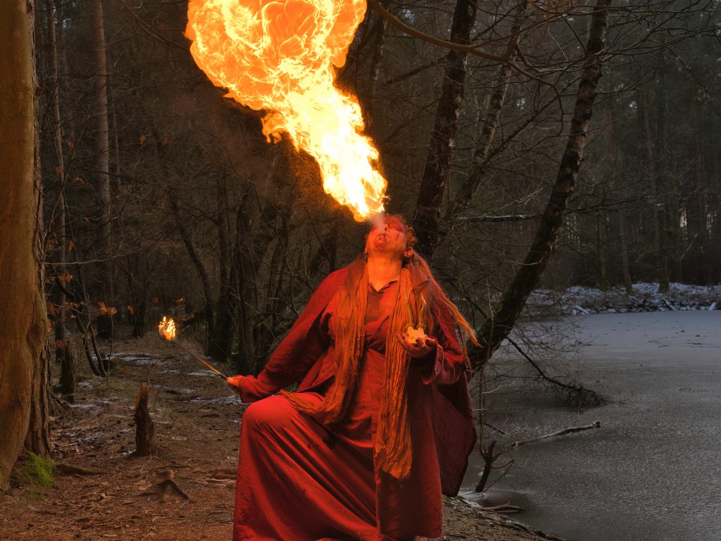 Fantasy-Portrait eines Feuerspuckers in rotem Gewand, der im Wald eine große Flamme erzeugt.