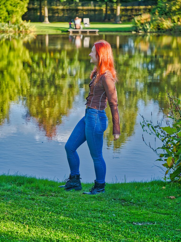Frau mit roten Haaren steht am Seeufer im Sonnenlicht, umgeben von grüner Natur und Wasserreflexionen.