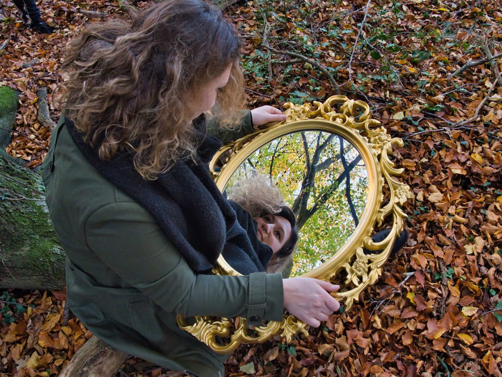 Frau mit lockigem Haar hält goldenen Spiegel im Herbstwald, in dem sich ihr Gesicht und Baumkronen spiegeln.