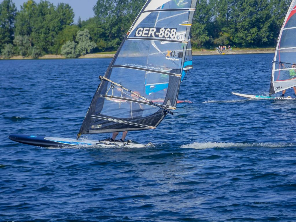 Zwei Windsurfer gleiten bei Sonnenschein über einen blauen See, im Hintergrund grüne Uferlandschaft.