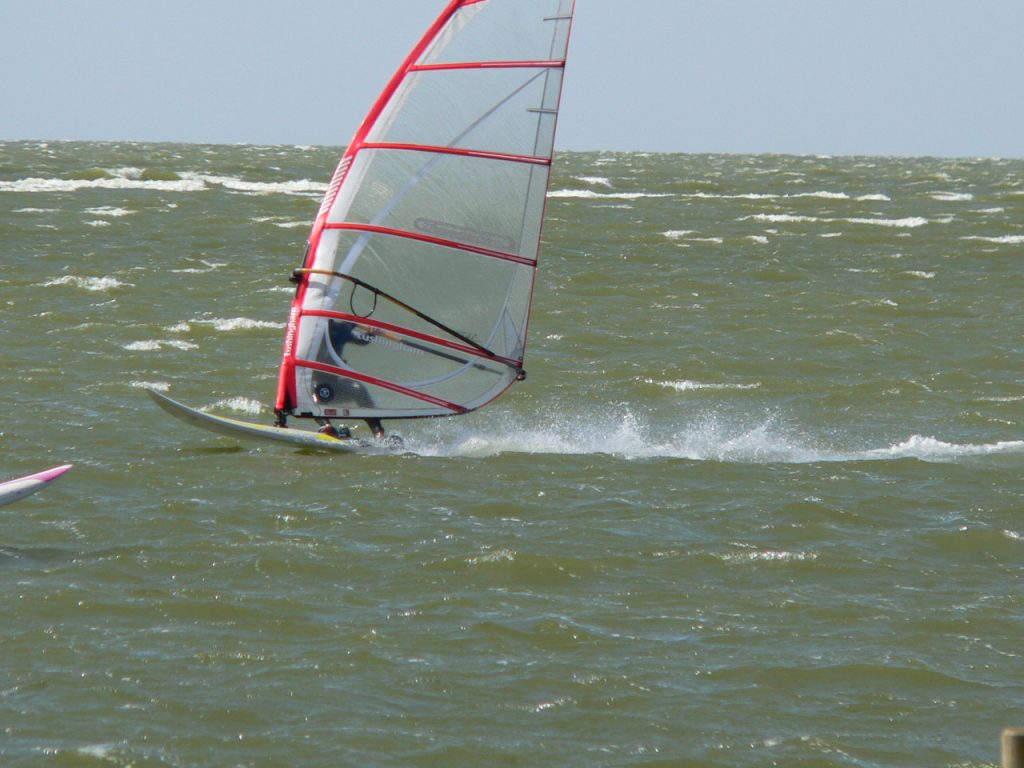 Windsurfer mit rotem Segel gleitet über wellige See bei starkem Wind.