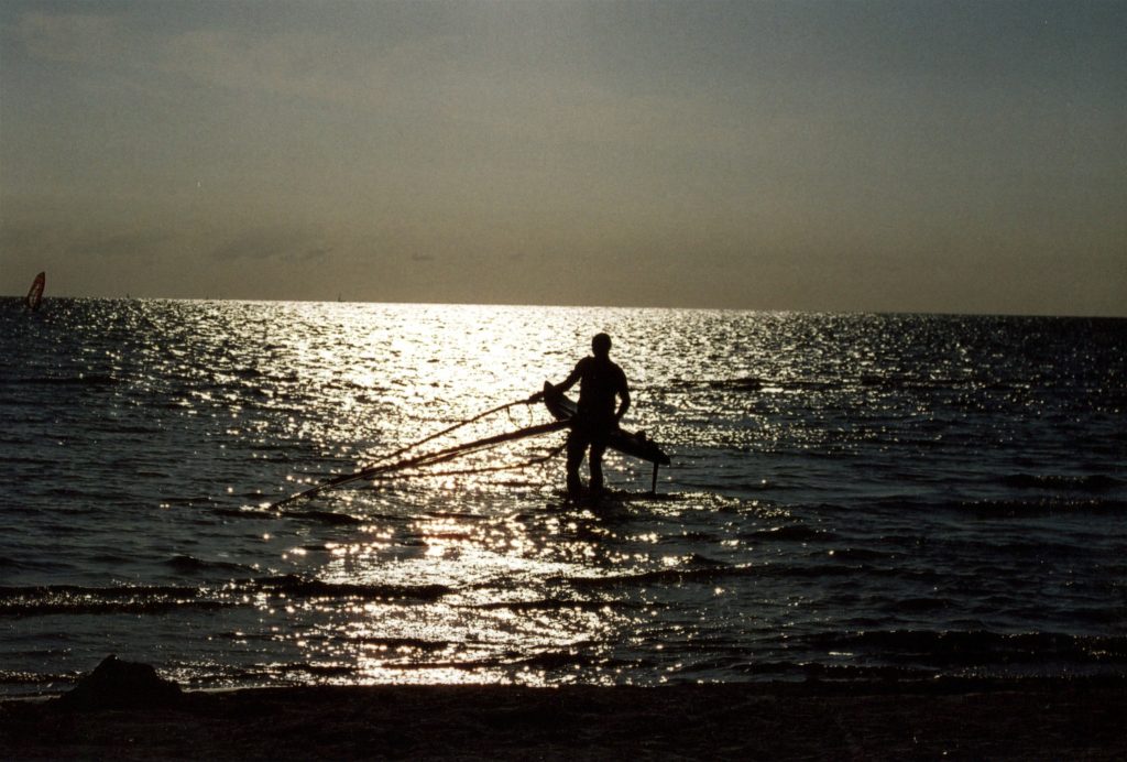 Silhouette eines Windsurfers im Meer bei Sonnenuntergang mit reflektierendem Wasser. Ein Beispiel für emotionale Sport Fotografie.