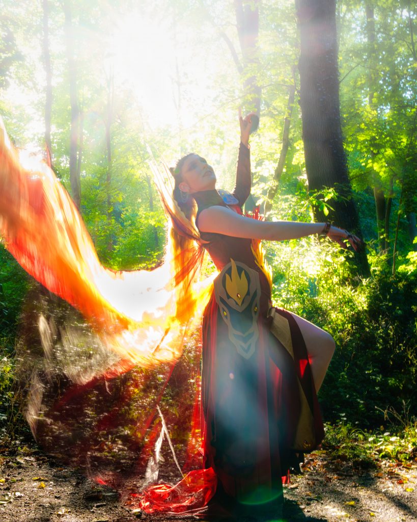 Fantasyfoto einer feuerfarbenen Cosplayerin mit leuchtenden Stoffen im Sonnenlicht.
