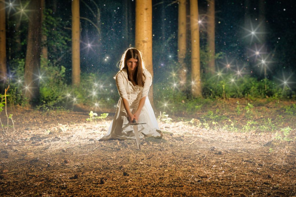 Fantasy Fotografie einer Frau in weißem Kleid mit Schwert im magisch leuchtenden Wald – kreative und atmosphärische Inszenierung mit Lichteffekten.