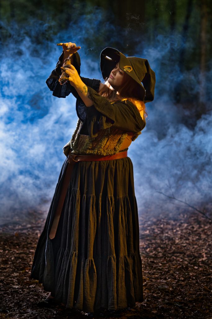 Fantasy-Fotografie einer Hexe im Wald, die ein kleines Objekt im aufsteigenden Rauch betrachtet