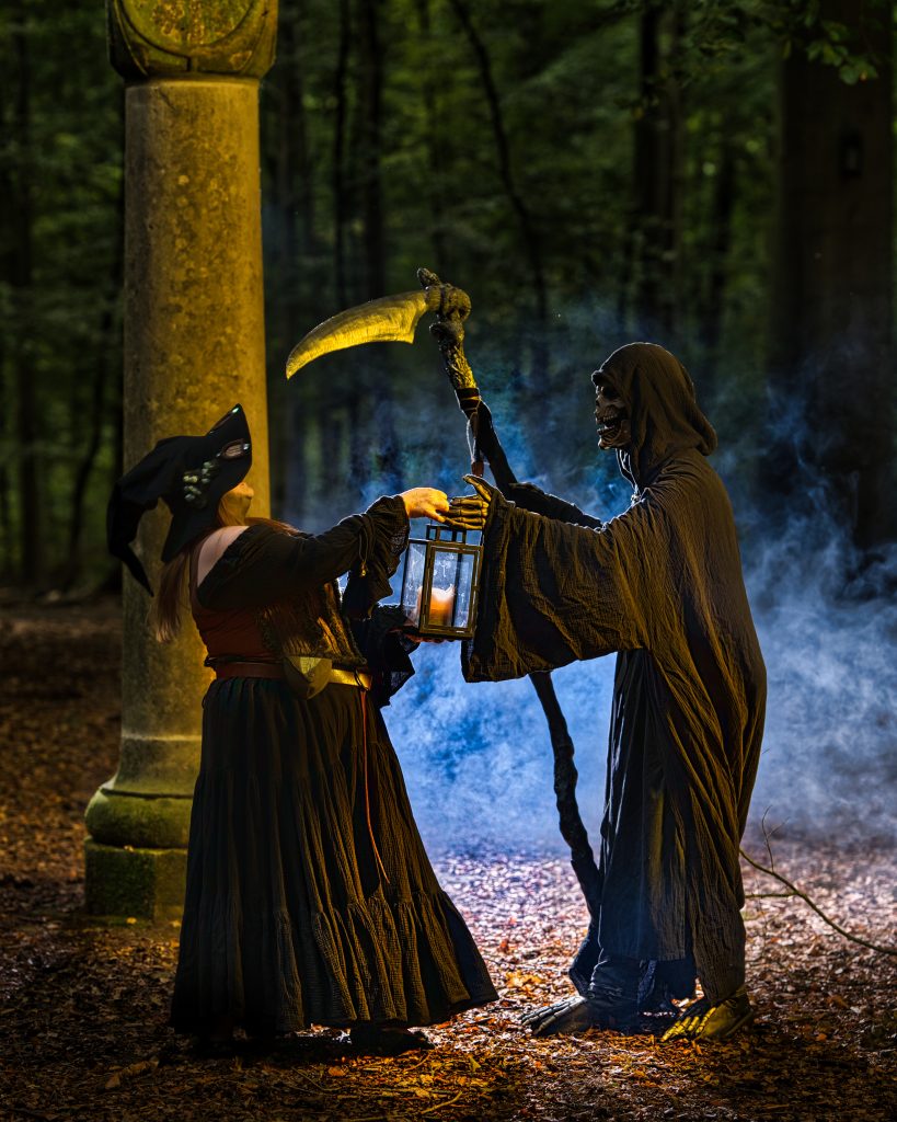 Fantasy-Fotografie einer Hexe im Wald, die einem Sensenmann mit Laterne an einer Steinsäule begegnet