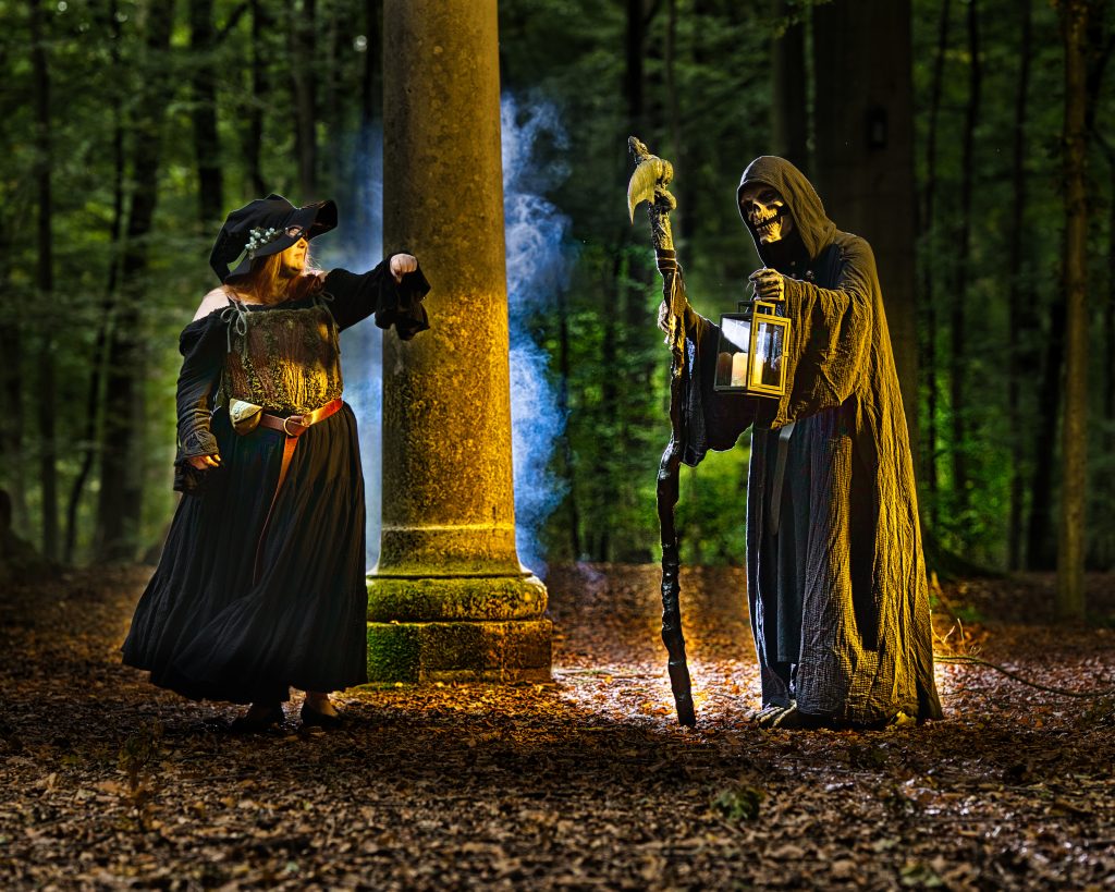 Fantasy-Szene im Wald mit zwei Figuren, eine mit Hut und Gewand, die andere als Sensenmann mit Laterne an einer Steinsäule