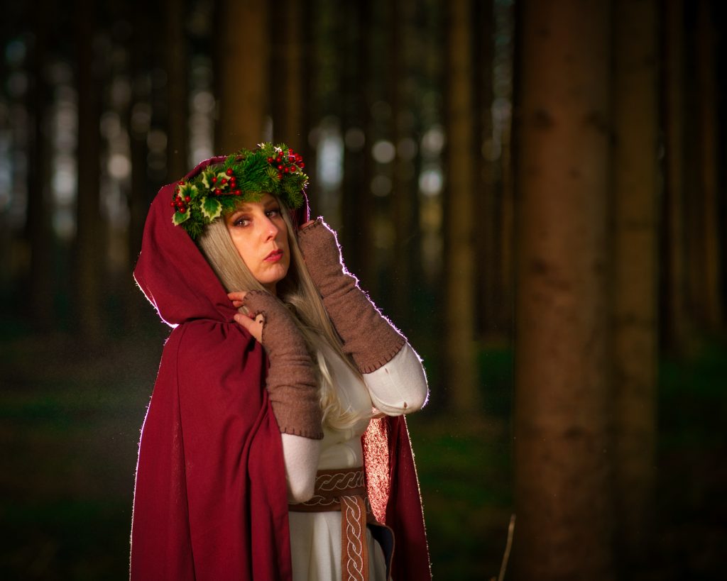 Frau in fantasy inspirierter Kleidung mit rotem Umhang und Tannenkranz, stehend im Wald bei gedämpftem Licht