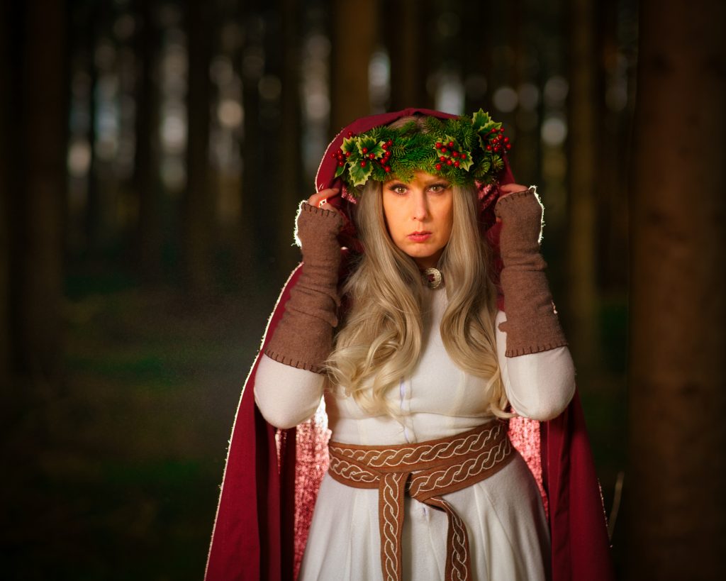 Frau in fantasyinspirierter Kleidung mit rotem Umhang und grünem Kranz aus Tannenzweigen und Beeren im Wald