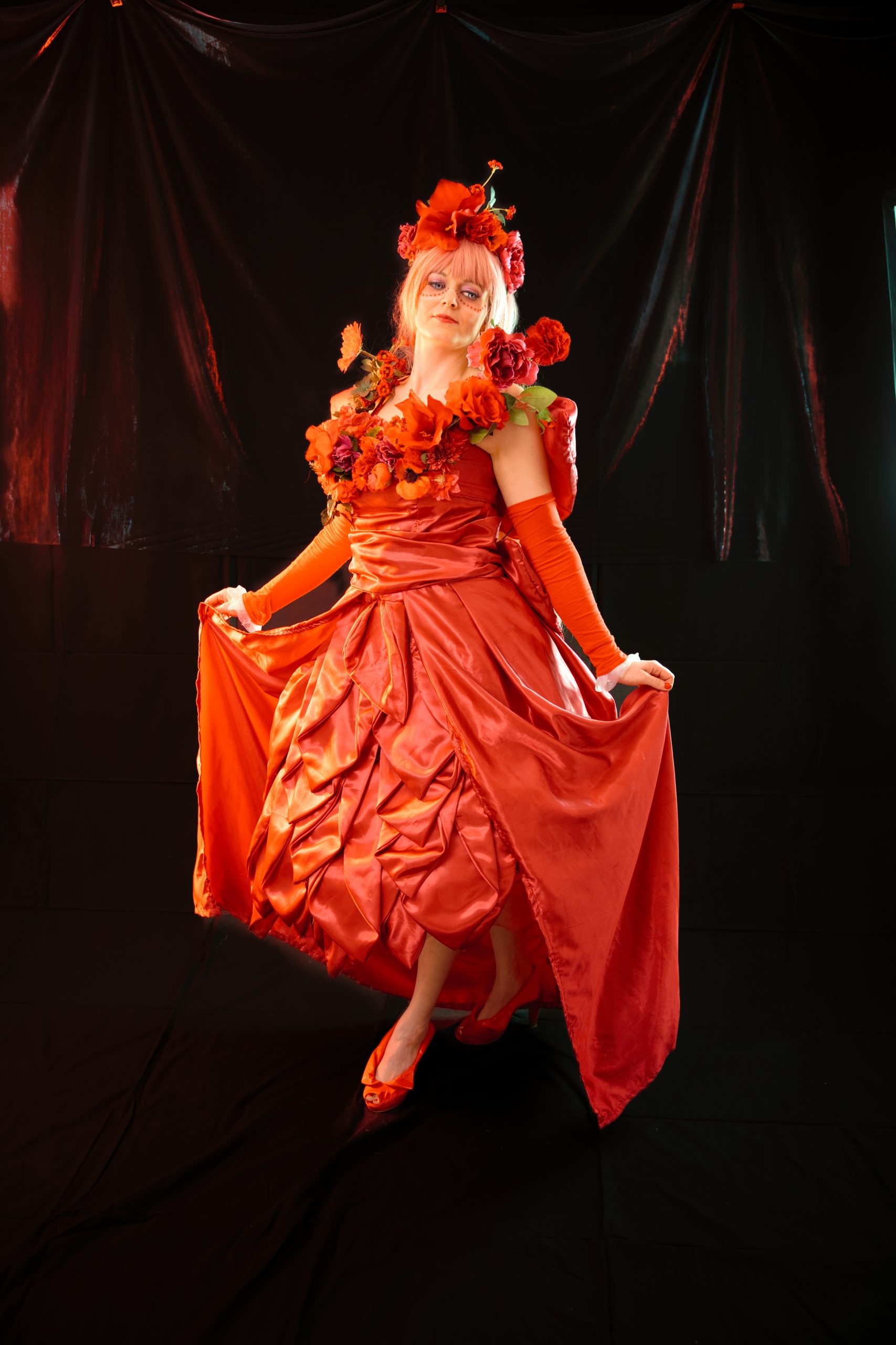 Cosplay-Aufnahme einer Person in rotem Kleid mit floralen Applikationen vor schwarzem Hintergrund