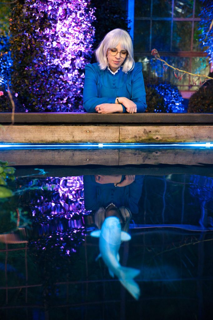 Fantasy-Fotografie einer Frau mit weißem Haar, die über einen beleuchteten Teich blickt, mit mystischer Spiegelung und Fisch im Wasser – Fantasy Fotografie von Thorsten Schwarz