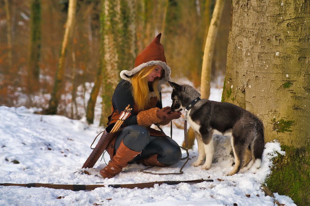 Fantasy-Fotografie einer Waldhüterin mit Kapuze, Lederkleidung und Husky im verschneiten Wald.