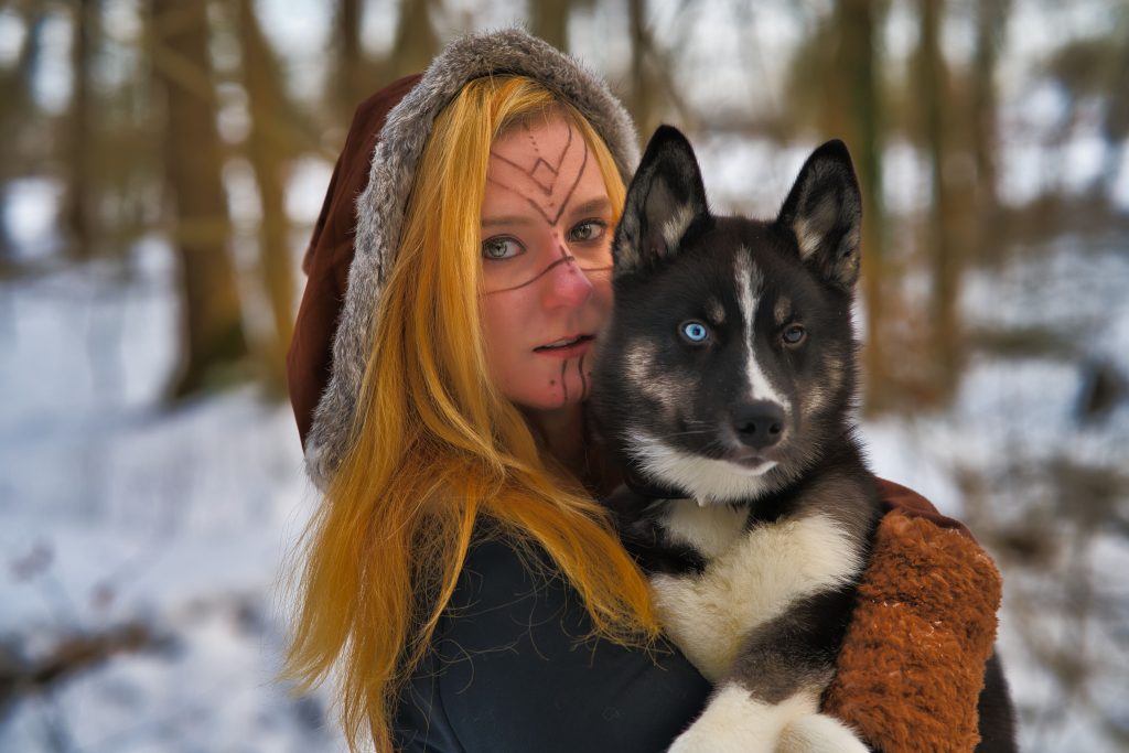 Fantasy-Porträt einer jungen Frau mit Runen-Gesichtsbemalung und Husky mit blauem Auge im winterlichen Wald – mystische Fotografie von Thorsten Schwarz