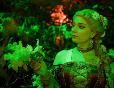 Fantasy-Fotografie einer jungen Frau im märchenhaften Kleid, die in grünem Licht eine Blüte im Wald betrachtet – stimmungsvolle Fantasy Fotografie von Thorsten Schwarz