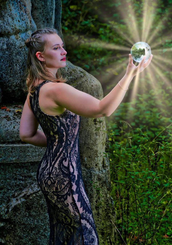 Fantasy Fotografie einer Frau in schwarzem Spitzenkleid, die eine leuchtende Kugel in der Natur hält.