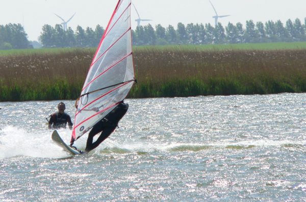 Windsurfer auf sonnigem Binnengewässer mit Windrädern und grüner Landschaft im Hintergrund.